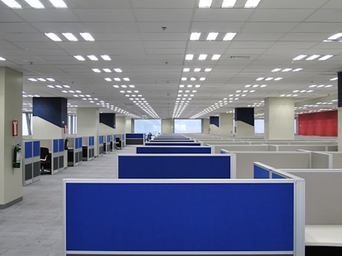 A large, empty office space with rows of blue cubicles. Overhead, fluorescent lights line the ceiling, and the room has a modern, orderly appearance. Fire extinguishers are visible on the walls, and windows at the far end allow natural light to filter in.