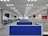 A large, empty office space with rows of blue cubicles. Overhead, fluorescent lights line the ceiling, and the room has a modern, orderly appearance. Fire extinguishers are visible on the walls, and windows at the far end allow natural light to filter in.