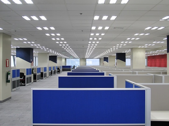 A large, empty office space with rows of blue cubicles. Overhead, fluorescent lights line the ceiling, and the room has a modern, orderly appearance. Fire extinguishers are visible on the walls, and windows at the far end allow natural light to filter in.
