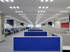 A large, empty office space with rows of blue cubicles. Overhead, fluorescent lights line the ceiling, and the room has a modern, orderly appearance. Fire extinguishers are visible on the walls, and windows at the far end allow natural light to filter in.