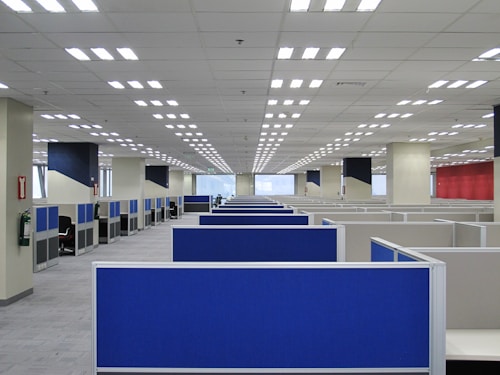 A large, empty office space with rows of blue cubicles. Overhead, fluorescent lights line the ceiling, and the room has a modern, orderly appearance. Fire extinguishers are visible on the walls, and windows at the far end allow natural light to filter in.