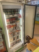 An open refrigerator with various packaged food items on display, including instant meals and canned goods. A person wearing a brown sweater and jeans is crouching next to the open door, possibly looking at the lower shelves.
