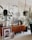 A cozy living room corner with a mid-century modern wooden sideboard featuring houseplants in various pots. Artworks are leaning against the cabinet, including a portrait and a family photo. A framed print with motivational text stands atop the sideboard, surrounded by stylish decor items such as a small lamp and a decorative container. The space is bright, with natural light coming through a window, and the overall decor exudes a homey and artistic vibe.