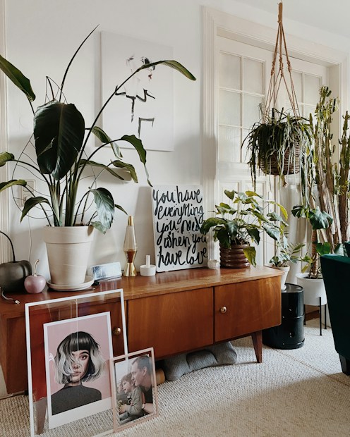 A cozy living room corner with a mid-century modern wooden sideboard featuring houseplants in various pots. Artworks are leaning against the cabinet, including a portrait and a family photo. A framed print with motivational text stands atop the sideboard, surrounded by stylish decor items such as a small lamp and a decorative container. The space is bright, with natural light coming through a window, and the overall decor exudes a homey and artistic vibe.