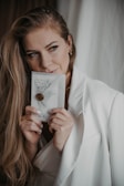 A person with long hair and a gentle expression holds a decorative envelope adorned with a wax seal. They are dressed in a light-colored blazer, and their subtle makeup enhances their thoughtful gaze.