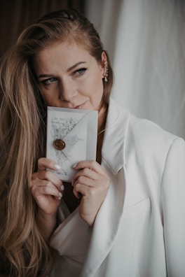 A person with long hair and a gentle expression holds a decorative envelope adorned with a wax seal. They are dressed in a light-colored blazer, and their subtle makeup enhances their thoughtful gaze.