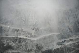 A barren and rugged landscape featuring a quarry or open-pit mine. Layers of excavated rock and earth form uneven, terraced levels. A narrow, winding road cuts through the scene, with small trucks or machinery visible, indicating industrial activity. The atmosphere appears dusty or foggy, softening the visibility of the scene.
