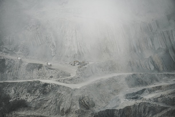 A barren and rugged landscape featuring a quarry or open-pit mine. Layers of excavated rock and earth form uneven, terraced levels. A narrow, winding road cuts through the scene, with small trucks or machinery visible, indicating industrial activity. The atmosphere appears dusty or foggy, softening the visibility of the scene.