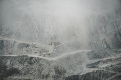 A barren and rugged landscape featuring a quarry or open-pit mine. Layers of excavated rock and earth form uneven, terraced levels. A narrow, winding road cuts through the scene, with small trucks or machinery visible, indicating industrial activity. The atmosphere appears dusty or foggy, softening the visibility of the scene.
