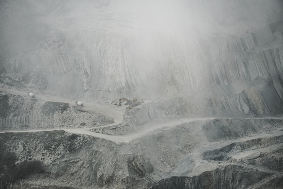 A barren and rugged landscape featuring a quarry or open-pit mine. Layers of excavated rock and earth form uneven, terraced levels. A narrow, winding road cuts through the scene, with small trucks or machinery visible, indicating industrial activity. The atmosphere appears dusty or foggy, softening the visibility of the scene.