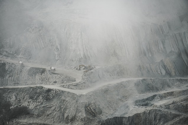 A barren and rugged landscape featuring a quarry or open-pit mine. Layers of excavated rock and earth form uneven, terraced levels. A narrow, winding road cuts through the scene, with small trucks or machinery visible, indicating industrial activity. The atmosphere appears dusty or foggy, softening the visibility of the scene.