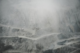 A barren and rugged landscape featuring a quarry or open-pit mine. Layers of excavated rock and earth form uneven, terraced levels. A narrow, winding road cuts through the scene, with small trucks or machinery visible, indicating industrial activity. The atmosphere appears dusty or foggy, softening the visibility of the scene.