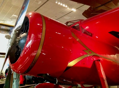A vibrant red aircraft engine with a silver propeller, accented by gold lines. The background includes a museum setting with visible overhead lighting and parts of other exhibits.