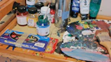 A rustic wooden table strewn with paintbrushes, jars of pigments, and a half-finished floral watercolor.