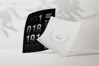 Two pieces of paper are arranged artistically on a plain surface. One is black with large numbers and text stating '1 000 000 views & counting.' The other is white with a minimal design featuring a small icon surrounded by laurels. Shadows of leaves are subtly cast on the background, adding depth.
