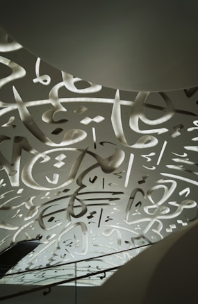 A ceiling design featuring intricate, abstract calligraphic patterns with illuminated cutouts. The patterns appear fluid and artistic, spreading across the entire surface. Gentle lighting enhances the visual appeal, creating a calming atmosphere.
