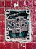 An exposed and weathered electrical socket set against a textured red tile background. The socket shows signs of rust and dirt, with visible wiring inside.