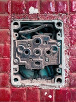 An exposed and weathered electrical socket set against a textured red tile background. The socket shows signs of rust and dirt, with visible wiring inside.