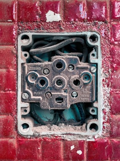 An exposed and weathered electrical socket set against a textured red tile background. The socket shows signs of rust and dirt, with visible wiring inside.