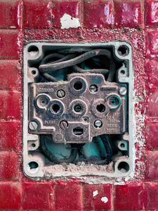 An exposed and weathered electrical socket set against a textured red tile background. The socket shows signs of rust and dirt, with visible wiring inside.
