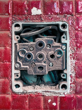 An exposed and weathered electrical socket set against a textured red tile background. The socket shows signs of rust and dirt, with visible wiring inside.