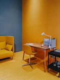 A cozy and modern office space featuring a wooden desk with a small white lamp and a few books and organizers. Next to it, there is a cushioned chair in a matching yellow shade. On the left, a mustard-yellow couch is accented with matching pillows. The floor and walls are painted in shades of yellow and blue, creating a warm and inviting atmosphere. To the right, a small black cart holds glasses and more books.