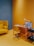 A cozy and modern office space featuring a wooden desk with a small white lamp and a few books and organizers. Next to it, there is a cushioned chair in a matching yellow shade. On the left, a mustard-yellow couch is accented with matching pillows. The floor and walls are painted in shades of yellow and blue, creating a warm and inviting atmosphere. To the right, a small black cart holds glasses and more books.
