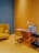 A cozy and modern office space featuring a wooden desk with a small white lamp and a few books and organizers. Next to it, there is a cushioned chair in a matching yellow shade. On the left, a mustard-yellow couch is accented with matching pillows. The floor and walls are painted in shades of yellow and blue, creating a warm and inviting atmosphere. To the right, a small black cart holds glasses and more books.