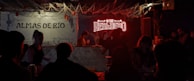 A dimly lit bar scene with a neon sign that reads 'HEREDERO' illuminating the area. Several people are gathered around the bar, with glasses and bottles visible on the counter. The left side has a wall sign 'ALMAS DE RÍO' and decorative elements like hanging ropes and small lights add to the ambiance.