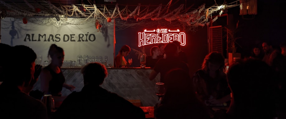 A dimly lit bar scene with a neon sign that reads 'HEREDERO' illuminating the area. Several people are gathered around the bar, with glasses and bottles visible on the counter. The left side has a wall sign 'ALMAS DE RÍO' and decorative elements like hanging ropes and small lights add to the ambiance.