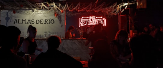 A dimly lit bar scene with a neon sign that reads 'HEREDERO' illuminating the area. Several people are gathered around the bar, with glasses and bottles visible on the counter. The left side has a wall sign 'ALMAS DE R&Iacute;O' and decorative elements like hanging ropes and small lights add to the ambiance.