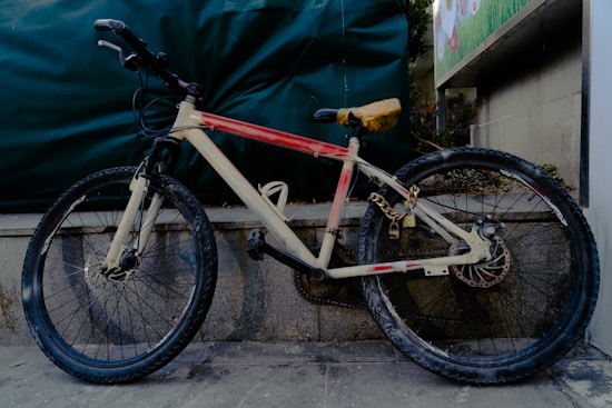 A bicycle with a beige and red frame is secured against a stone wall with a sturdy chain and padlock. The tires are worn, suggesting frequent use, and the seat is covered with a protective yellow cover. There is a green tarpaulin background and part of a colorful poster visible on the right.