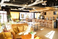 A cozy café interior featuring comfortable seating with plush sofas and wooden chairs. A long bar with high stools lines one side, and colorful artwork decorates the walls. Natural light filters through large windows with yellow curtains, and greenery adds a touch of nature. Industrial elements are visible in the exposed ductwork above.