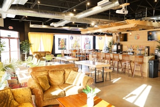 A cozy café interior featuring comfortable seating with plush sofas and wooden chairs. A long bar with high stools lines one side, and colorful artwork decorates the walls. Natural light filters through large windows with yellow curtains, and greenery adds a touch of nature. Industrial elements are visible in the exposed ductwork above.
