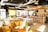 A cozy café interior featuring comfortable seating with plush sofas and wooden chairs. A long bar with high stools lines one side, and colorful artwork decorates the walls. Natural light filters through large windows with yellow curtains, and greenery adds a touch of nature. Industrial elements are visible in the exposed ductwork above.