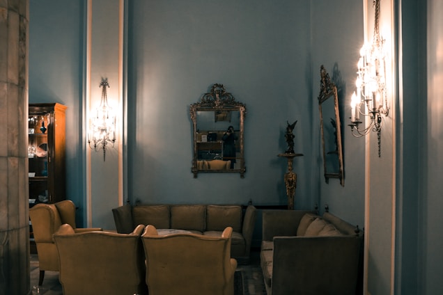 An elegant room furnished with vintage-style sofas and armchairs in a cozy arrangement. The walls are adorned with ornate mirrors and classic wall sconces, casting a warm glow. A display cabinet full of items is visible to the side, adding to the overall sophistication.