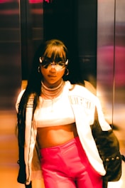 A stylish person wearing a cropped top, a trendy jacket with the word 'strength' visible, and vibrant pink pants. The individual is accessorized with sunglasses and layered necklaces, standing in what seems to be an elevator with metallic walls, under warm lighting that casts a golden hue.
