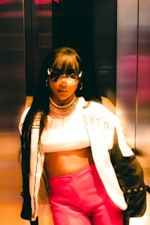 A stylish person wearing a cropped top, a trendy jacket with the word 'strength' visible, and vibrant pink pants. The individual is accessorized with sunglasses and layered necklaces, standing in what seems to be an elevator with metallic walls, under warm lighting that casts a golden hue.