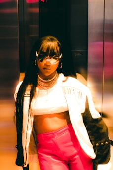 A stylish person wearing a cropped top, a trendy jacket with the word 'strength' visible, and vibrant pink pants. The individual is accessorized with sunglasses and layered necklaces, standing in what seems to be an elevator with metallic walls, under warm lighting that casts a golden hue.