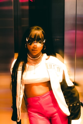 A stylish person wearing a cropped top, a trendy jacket with the word 'strength' visible, and vibrant pink pants. The individual is accessorized with sunglasses and layered necklaces, standing in what seems to be an elevator with metallic walls, under warm lighting that casts a golden hue.