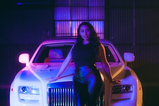 A woman with long, wavy hair stands confidently in front of a luxury white car with distinctive headlights and a prominent grille. The setting has an atmospheric lighting with purple and blue hues, enhancing the stylish and modern ambiance.