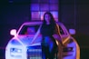 A woman with long, wavy hair stands confidently in front of a luxury white car with distinctive headlights and a prominent grille. The setting has an atmospheric lighting with purple and blue hues, enhancing the stylish and modern ambiance.