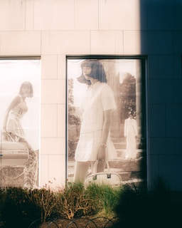 A large window display features a poster of a woman with short dark hair wearing a short-sleeved dress and holding a handbag. The background appears to depict a stone staircase and foliage, suggesting an outdoor setting. Another image of a woman, also with short hair and wearing a patterned dress, is seen partially in another window. The scene has a vintage and artistic feel, further emphasized by the soft lighting and shadows cast on the building facade.
