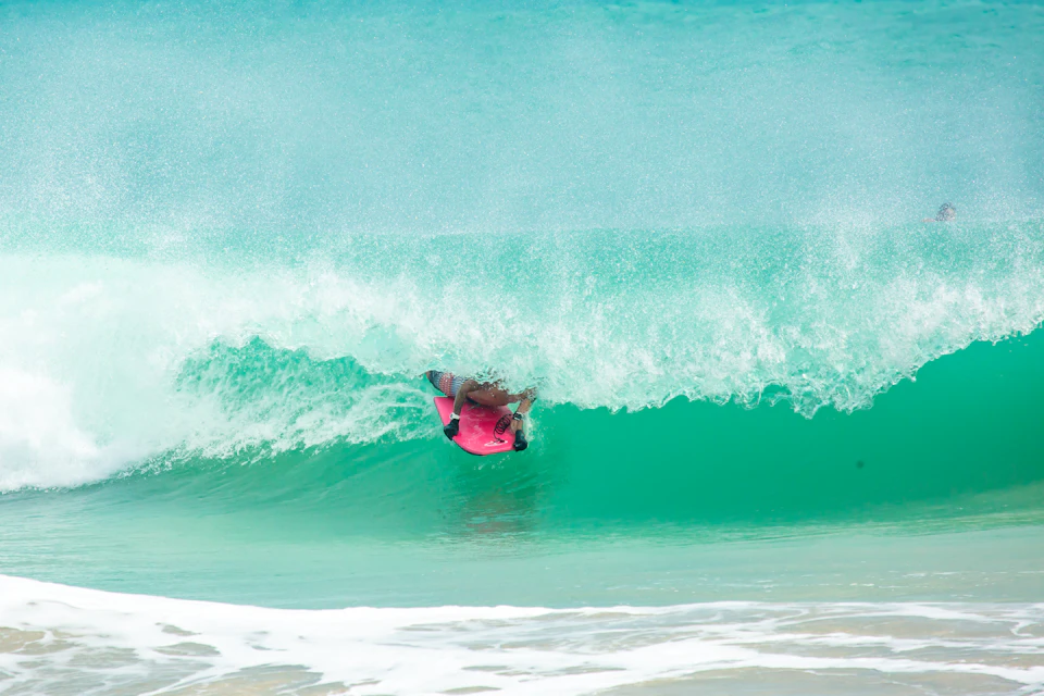 Surfer rides pink surfboard through turquoise-green ocean wave with white foam and blurred bystanders