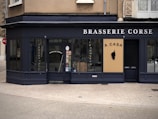A corner restaurant with a dark exterior features the name 'Brasserie Corse' prominently displayed. The main entrance door has the words 'Entrée des Artistes' on it. A beige sign with black text 'A CASA' and a silhouette of a head emblem is prominently displayed. Large windows showcase a glimpse of the interior.