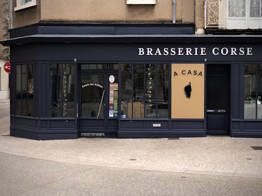A corner restaurant with a dark exterior features the name 'Brasserie Corse' prominently displayed. The main entrance door has the words 'Entrée des Artistes' on it. A beige sign with black text 'A CASA' and a silhouette of a head emblem is prominently displayed. Large windows showcase a glimpse of the interior.