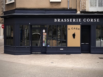 A corner restaurant with a dark exterior features the name 'Brasserie Corse' prominently displayed. The main entrance door has the words 'Entrée des Artistes' on it. A beige sign with black text 'A CASA' and a silhouette of a head emblem is prominently displayed. Large windows showcase a glimpse of the interior.