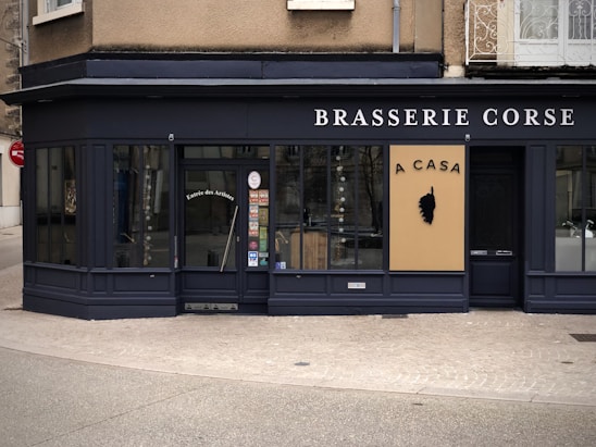A corner restaurant with a dark exterior features the name 'Brasserie Corse' prominently displayed. The main entrance door has the words 'Entrée des Artistes' on it. A beige sign with black text 'A CASA' and a silhouette of a head emblem is prominently displayed. Large windows showcase a glimpse of the interior.