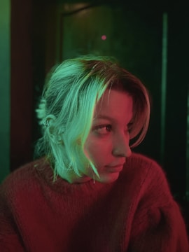 A person with light hair is illuminated by colored lighting, creating a dramatic and moody atmosphere. The lighting casts green and red hues on the person's face and sweater, adding a unique artistic effect.