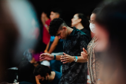 Several people appear to be in a gathering, some with their heads bowed and their eyes closed, suggesting a moment of concentration or reflection. One person is wearing a face mask while another is in focus with patterned clothing.